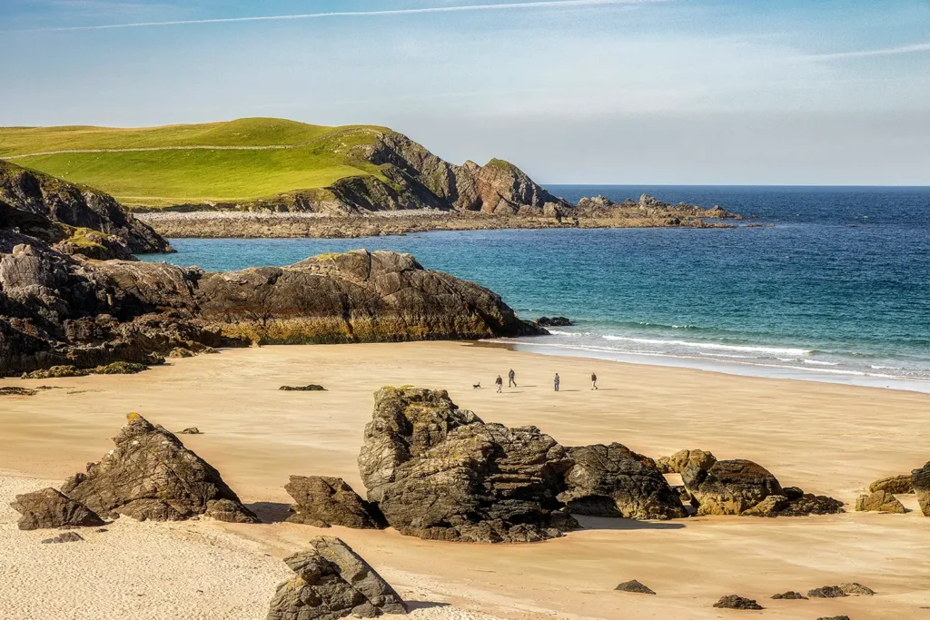 Plage de sable clair bordée de rochers et de collines verdoyantes, face aux eaux turquoise de la mer sur la côte écossaise