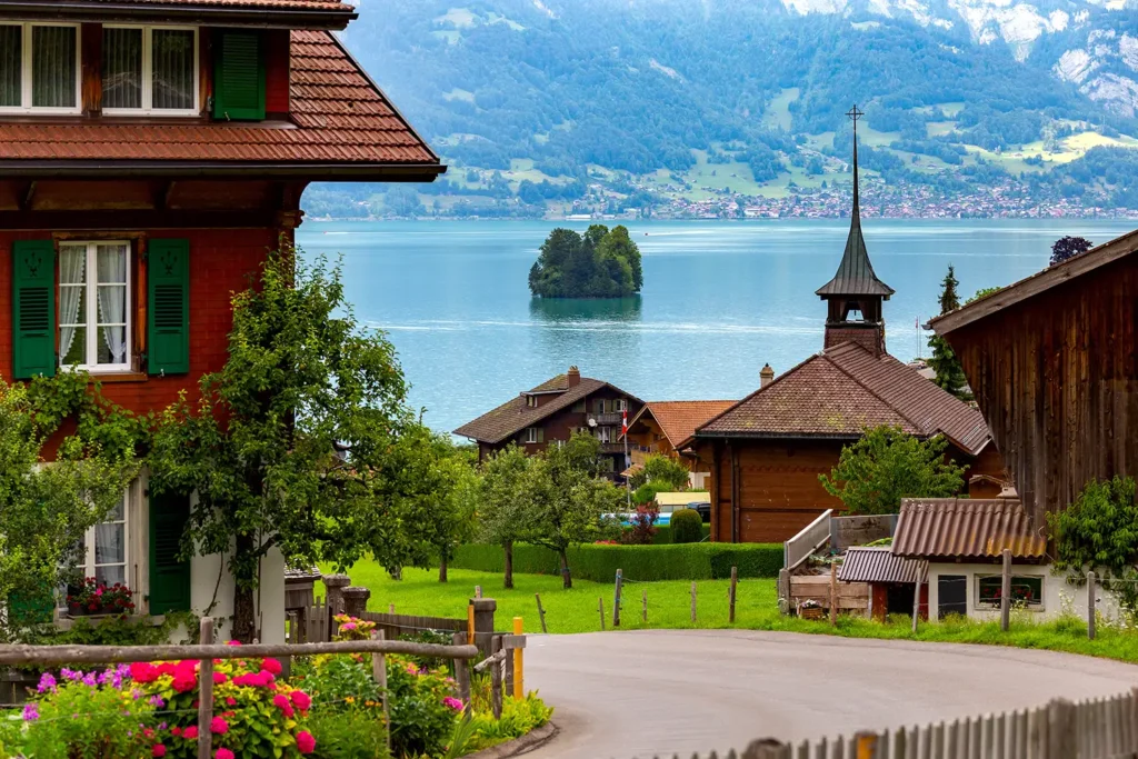 Chalets traditionnels en bois et petite église surplombant un lac suisse, avec un îlot boisé et des montagnes alpines en arrière-plan