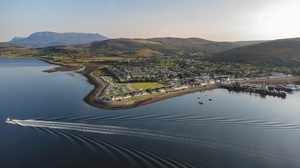 Vue aérienne d’un village portuaire écossais, avec ses maisons alignées au bord d’un loch, un port, des bateaux et des collines en arrière-plan